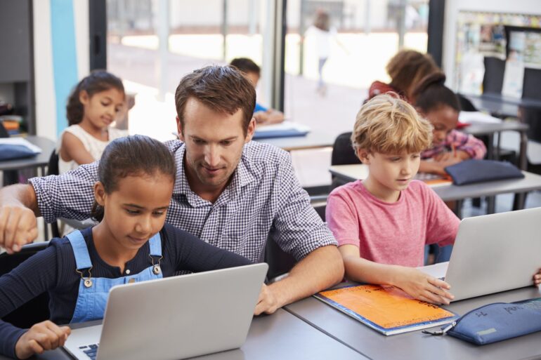 Teacher instructing young students who are using laptops.