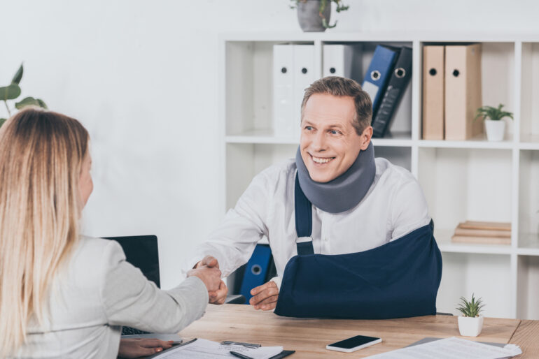 blond woman shaking hands with worker in neck brace and arm bandage over table in office, compensation concept