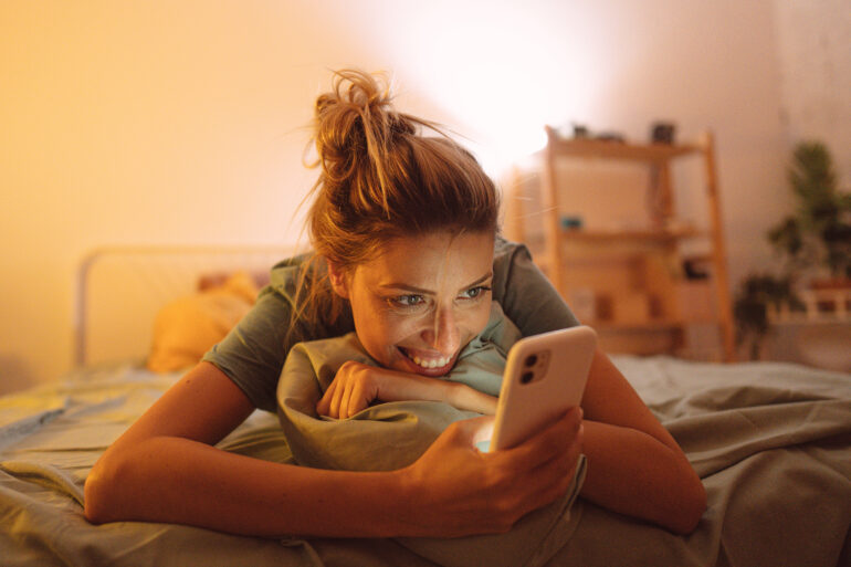 Photo of a young, smiling woman has a video call at night while lying on a bed in her bedroom